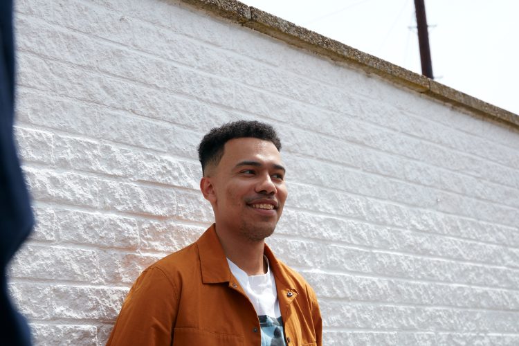 Josh stood against a painted white brick wall, smiling and looking away from camera. He's wearing a white graphic t-shirt with a mustard coloured jacket over