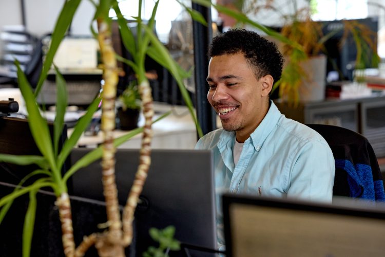 Josh sat at his desk smiling whilst working. In the foreground is a plant and computer screens