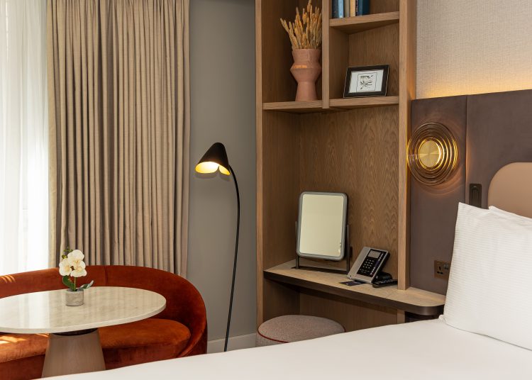 A hotel bedroom with a glimpse of the bed in the foreground with open wooden shelving to the left. There's a coffee table and amber coloured sofa in front of a tall window.