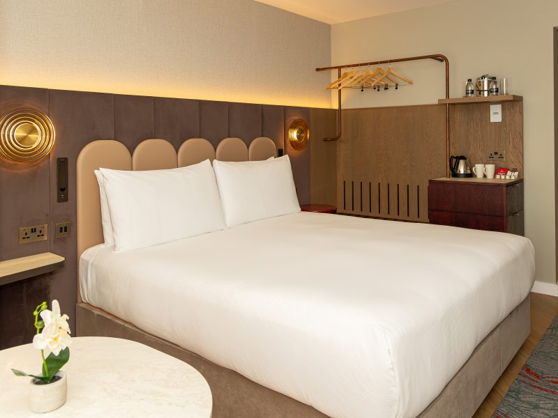 A hotel bedroom featuring a large bed with white bedding and a scalloped headboard, modern brown panels on the walls and an open clothes hanging rail.