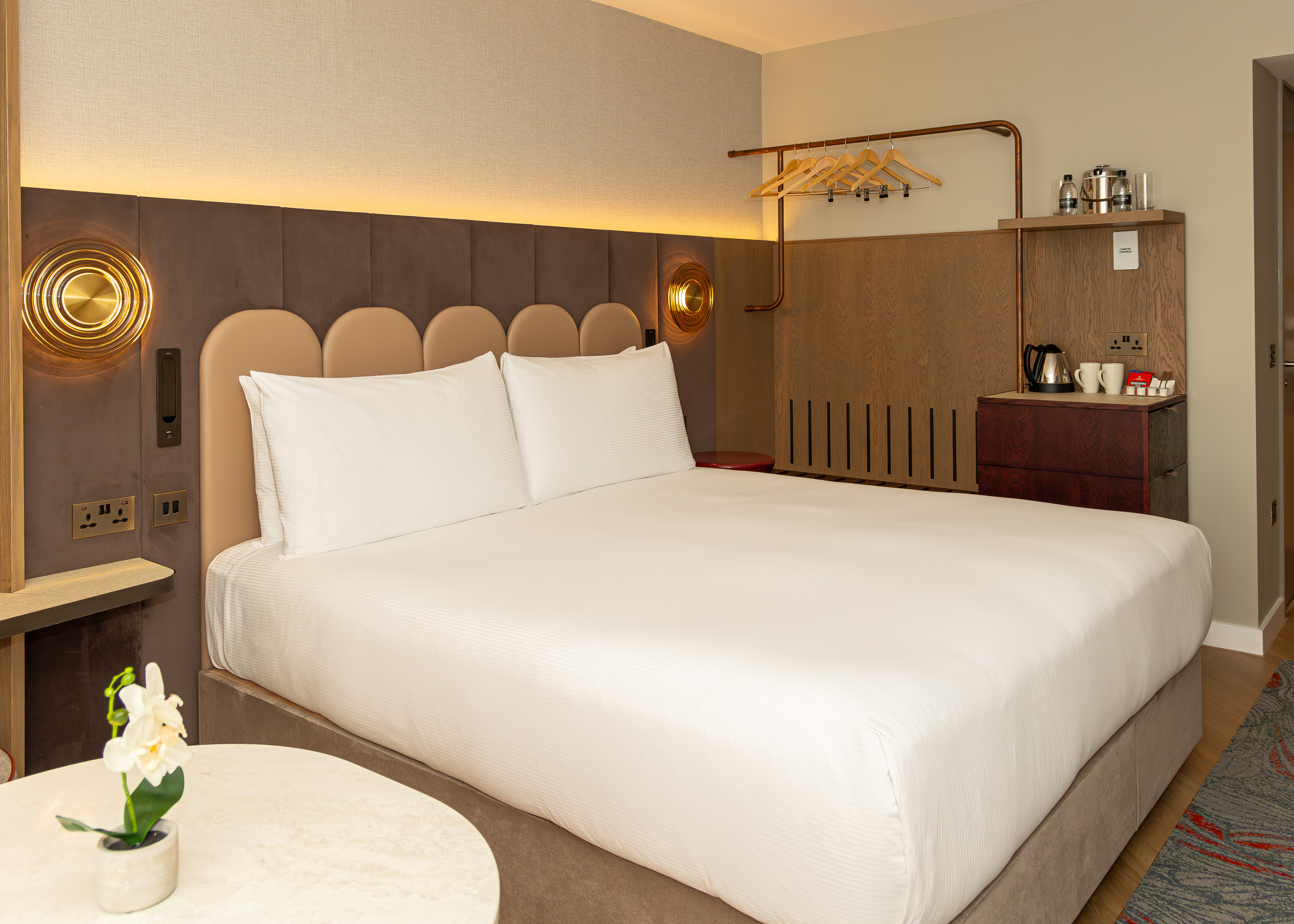 A hotel bedroom featuring a large bed with white bedding and a scalloped headboard, modern brown panels on the walls and an open clothes hanging rail.
