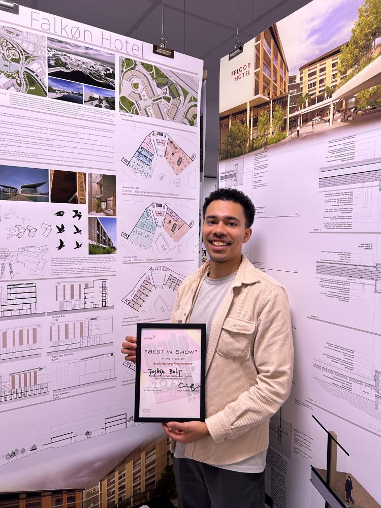 Josh proudly holding his framed award, stood in front of his university display.