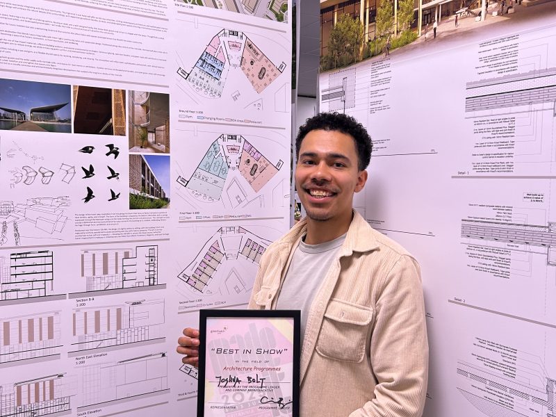 Josh proudly holding his framed award, stood in front of his university display.