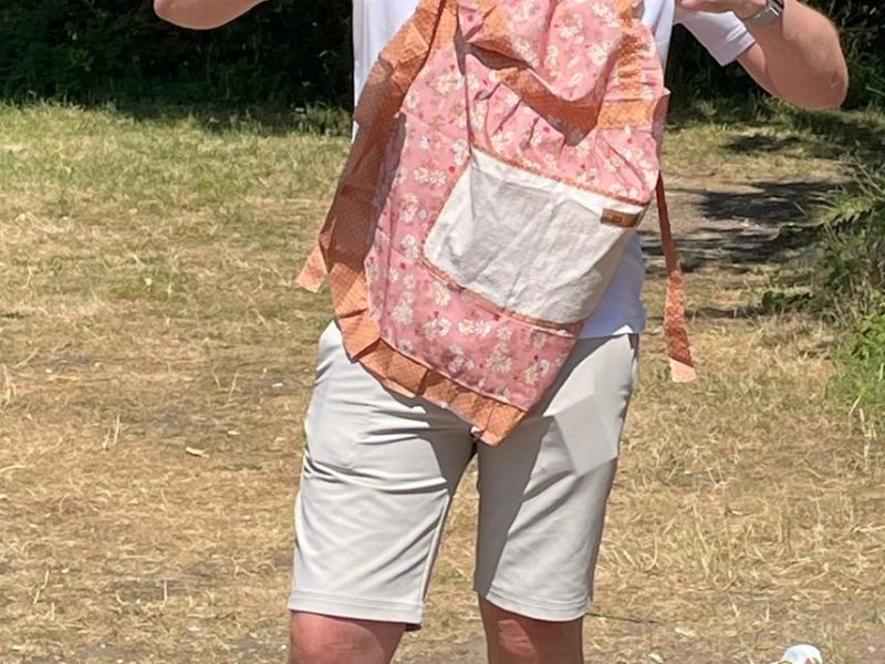 Peter Newman_Earp wearing light coloured shorts and a t shirt together with a pink apron. He's standing outside on a grassy area with trees behind him and he's giving a thumbs up.