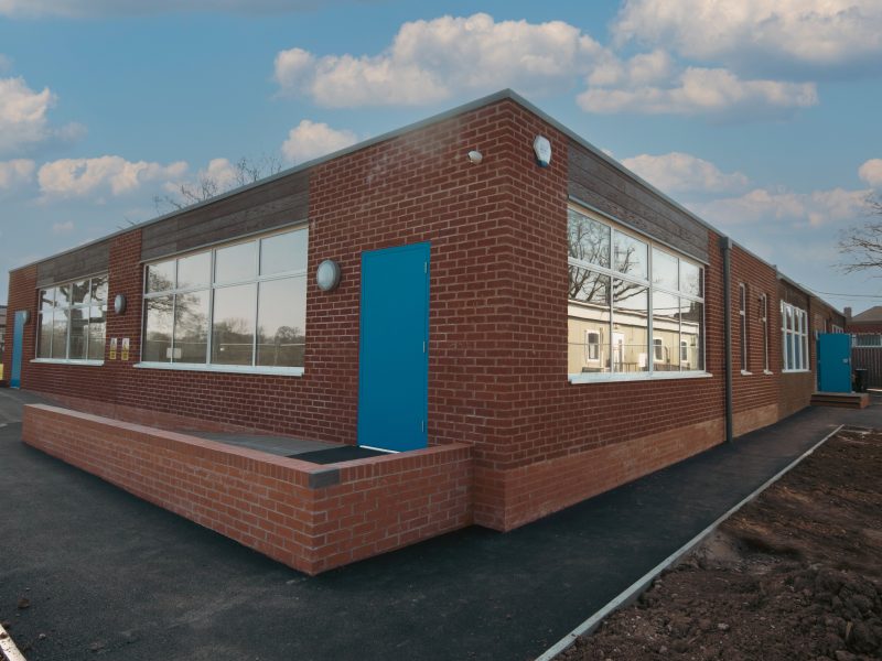 A modern, single-story brick building with large windows and a bright blue door, surrounded by a paved walkway under a partly cloudy sky.