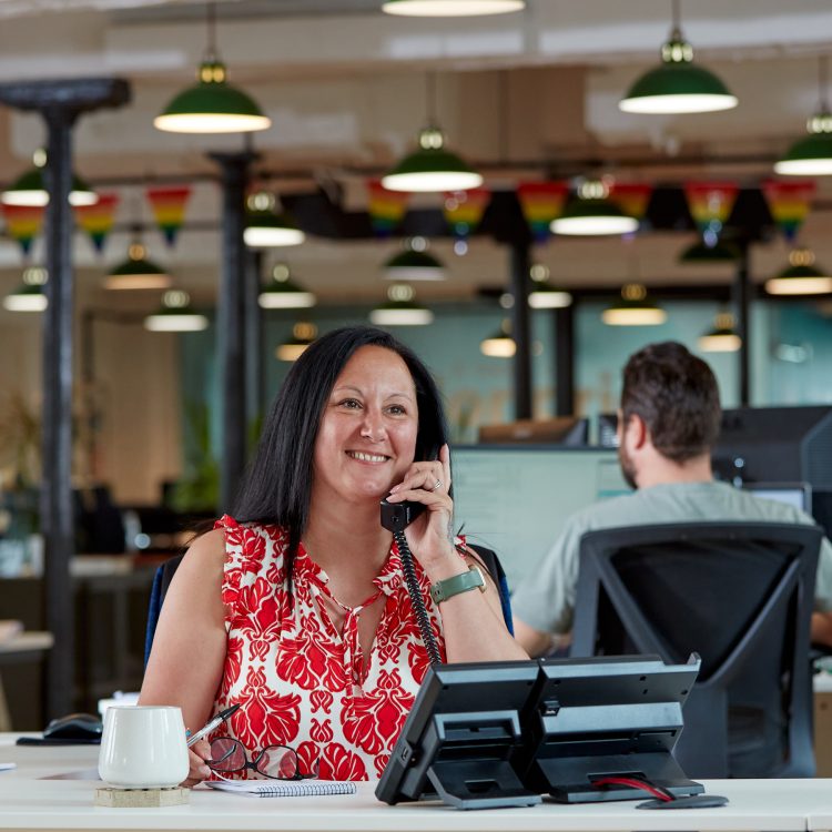 Image of Jo sitting at her desk in the studio. She's wearing a red and white patterned top and is on the phone.