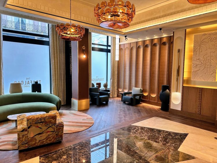 The Trafalgar St. James Hotel reception area. Featuring a mixture of wood and marble flooring, large windows, a green curved sofa, panelled walls and statement pendant lights.