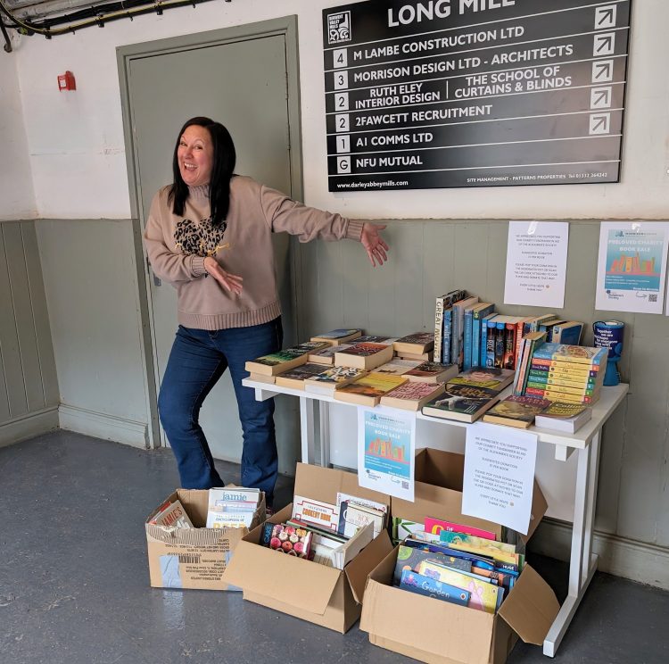 Jo is in the foyer of Darley Abbey Mills showing off our charity book sale which is set up on a table. She's wearing dark blue jeans and a beige jumper with a leopard print heart.