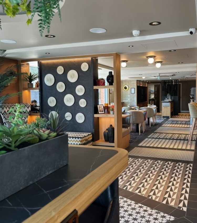 A photograph through to the breakfast area inside the hotel. It has monochrome, patterned tile flooring with tables and chairs set up. In front of the seating area is a large shelved partition featuring accessories and a dark panel in the centre with a pale circle design.