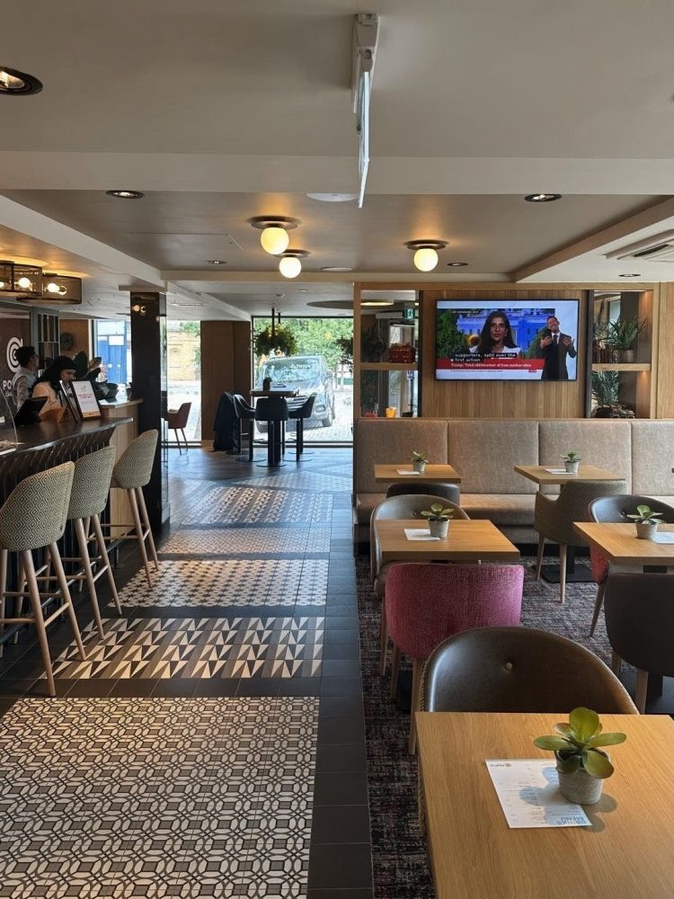 A photograph of the breakfast area inside the hotel with the bar on the left. It has monochrome, patterned tile flooring with tables and chairs set up on the right hand side. There is a TV mounted on a large shelved partition at the back of the seating area and you can see the glazed entrance through to the back of the photo.