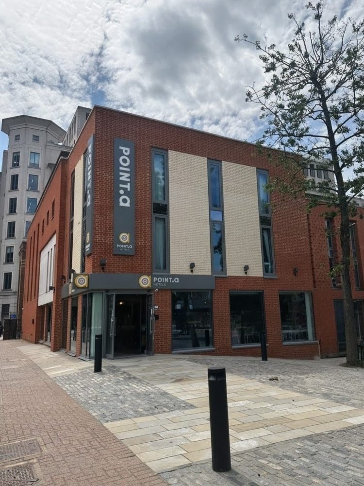 A photograph of the Point A Canary Wharf building from the street. A large red brick building with the entrance at the corner. It has large, grey Point A signage high on each corner of the building and around the entrance. There are three large windows to the right of the entrance and some narrow ones above.