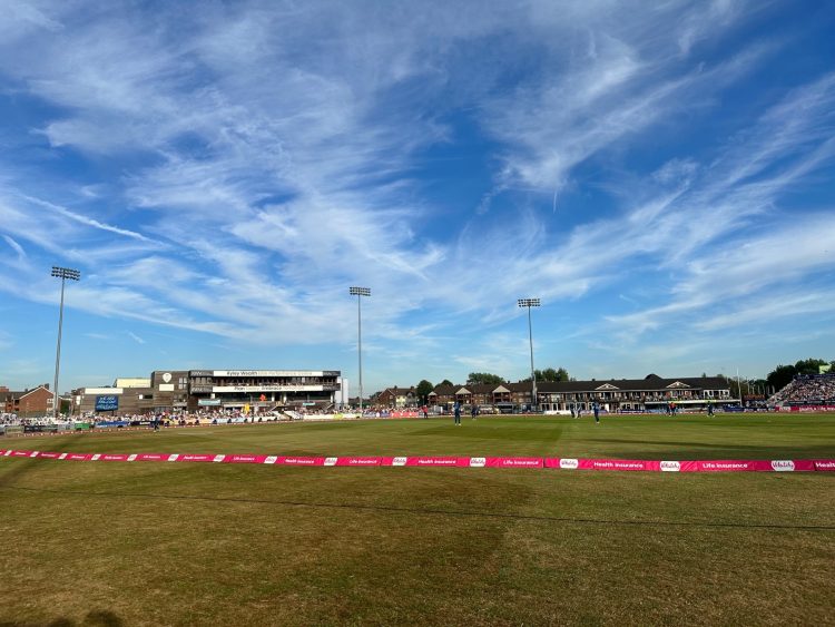 The view of the cricket pitch on a sunny day with stands and the crowd on the far side of the pitch.