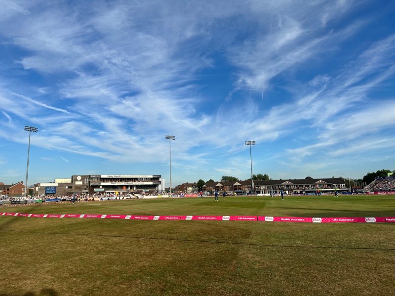 The view of the cricket pitch on a sunny day with stands and the crowd on the far side of the pitch.