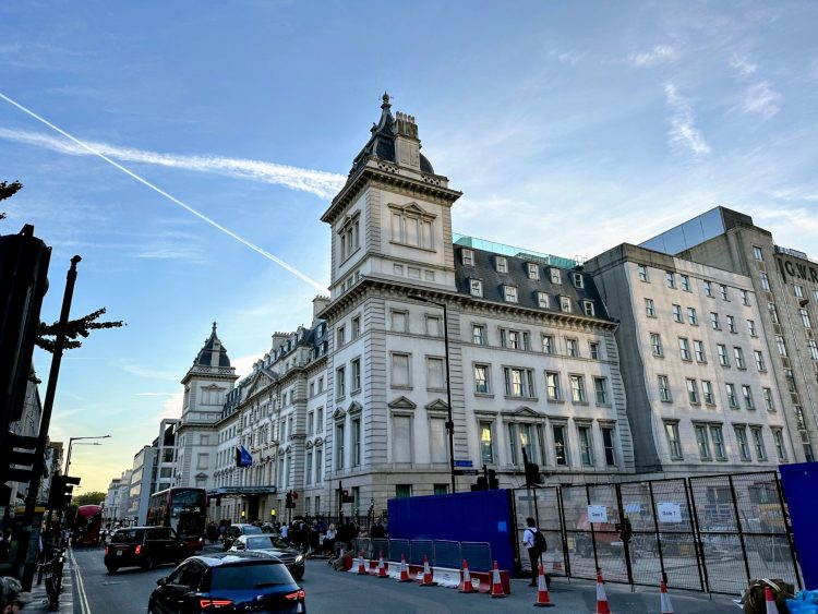 An external view of Hilton Paddington - a tall building with a prominent towers. It’s surrounded by a fence where construction is underway, located in an urban area with vehicles parked on the road and a blue sky filled with clouds above.