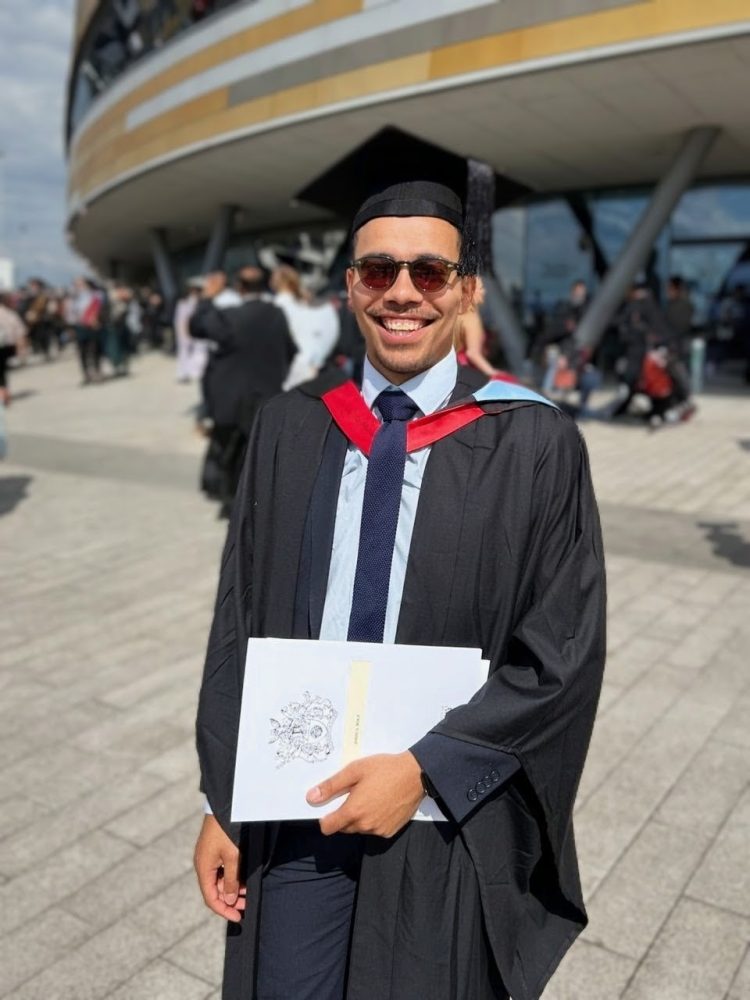 Josh standing outside the University of Derby, wearing his cap and gown and proudly holding his degree papers.