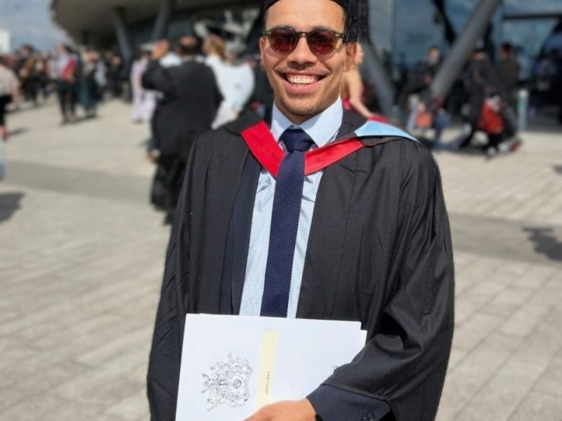 Josh standing outside the University of Derby, wearing his cap and gown and proudly holding his degree papers.