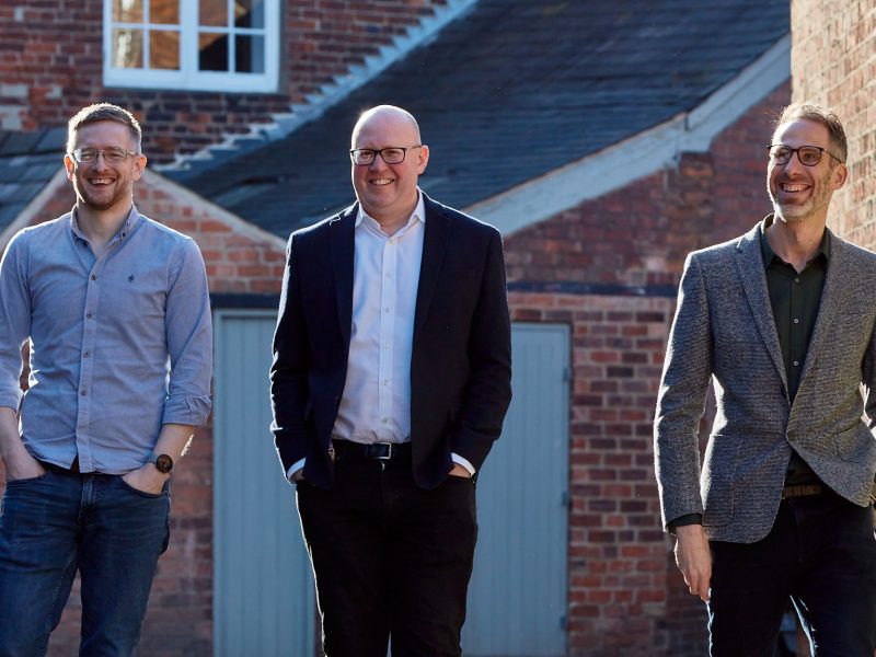 Directors David Gardner, Peter Newman-Earp and Nik Clarke walking together with the red brick mill buildings in the background.