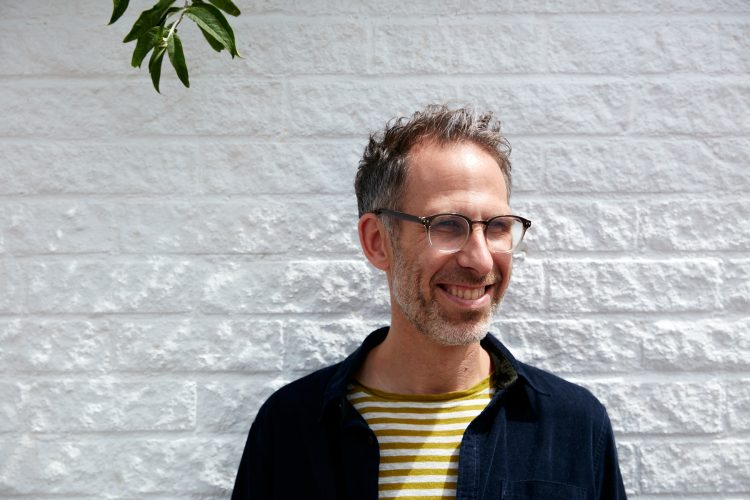 Nik standing in front of a white brick wall. He's smiling and wearing a green and white stripe top with a dark navy jacket over.
