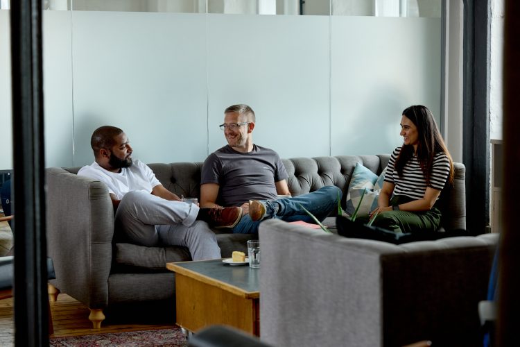 Two of our Diversity & Inclusion team, Sef and Nidhi, chatting with our Director David Gardner. They're sat on a grey sofa, smiling.