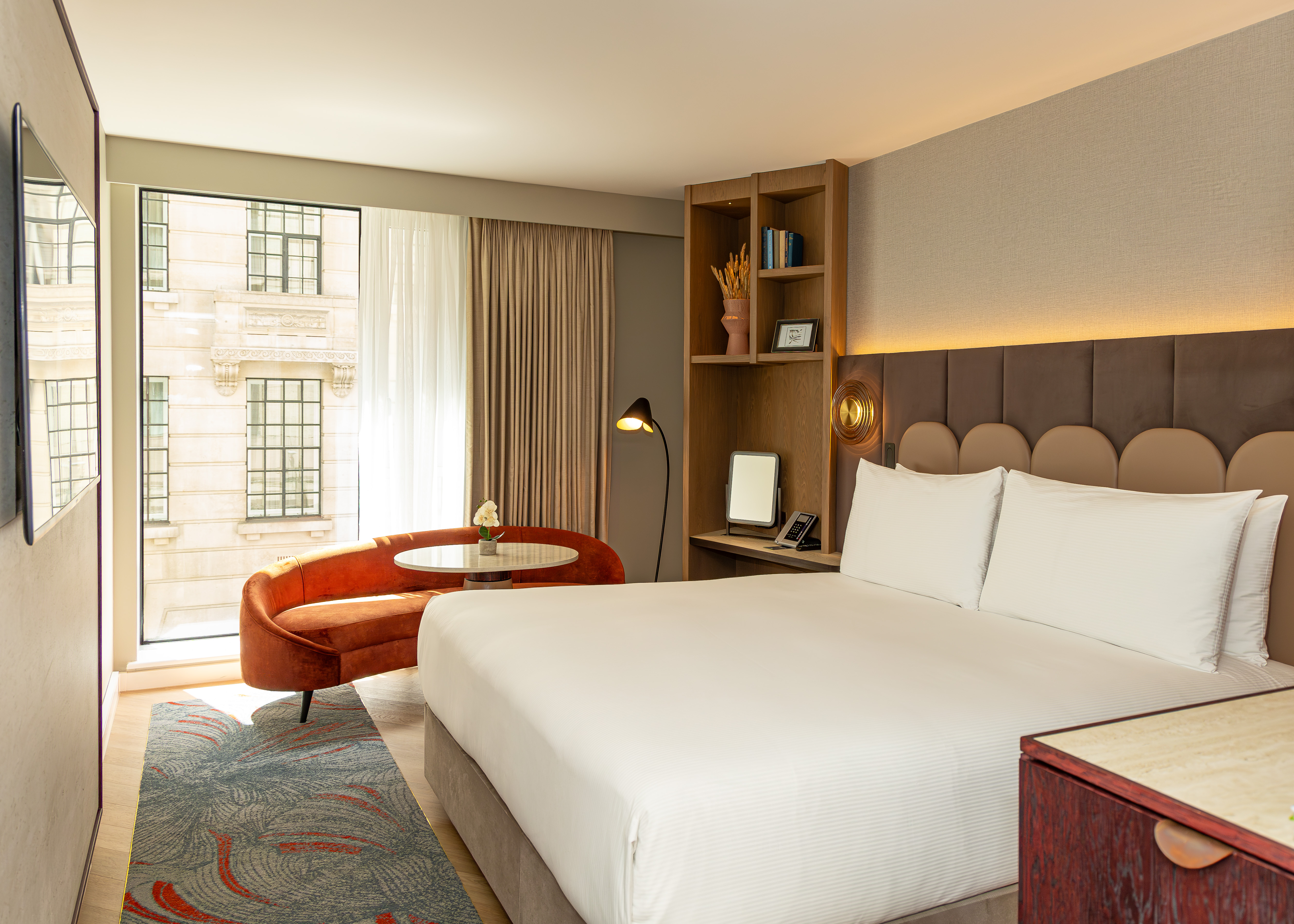Image of a newly refurbished guest bedroom taken from the doorway. There’s a full height window behind a burnt orange sofa and white coffee table. The bed has crisp white bedding and scalloped headboard in front of luxurious panels. There’s a wooden shelving unit in the corner of the room.