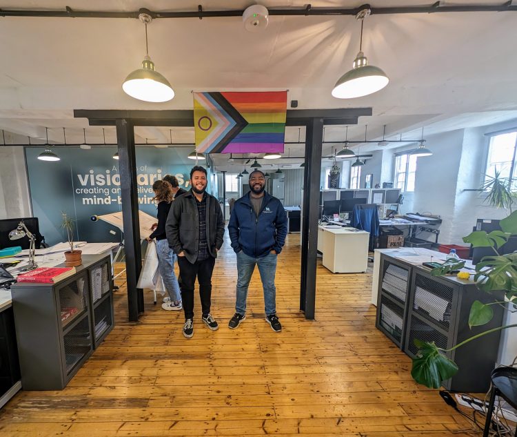 Sunny Kochchat and Sef Lawrence standing in the studio underneath a Pride flag.