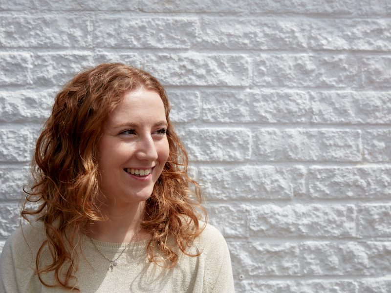 Photograph of Sophie stood in front of a painted white brick wall. She's smiling and looking away from camera, wearing a cream knit jumper.