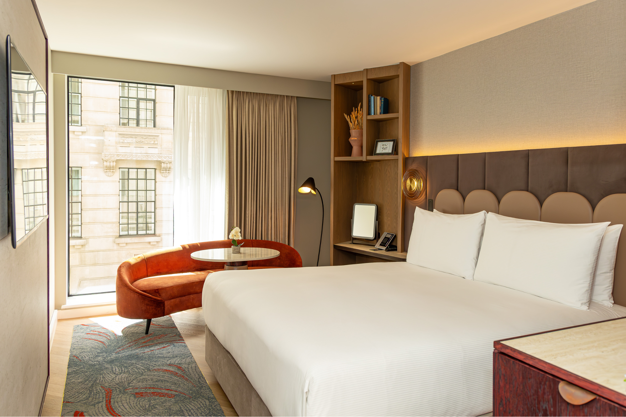 Image of a newly refurbished guest bedroom taken from the doorway. There’s a full height window behind a burnt orange sofa and white coffee table. The bed has crisp white bedding and scalloped headboard in front of luxurious panels. There’s a wooden shelving unit in the corner of the room.
