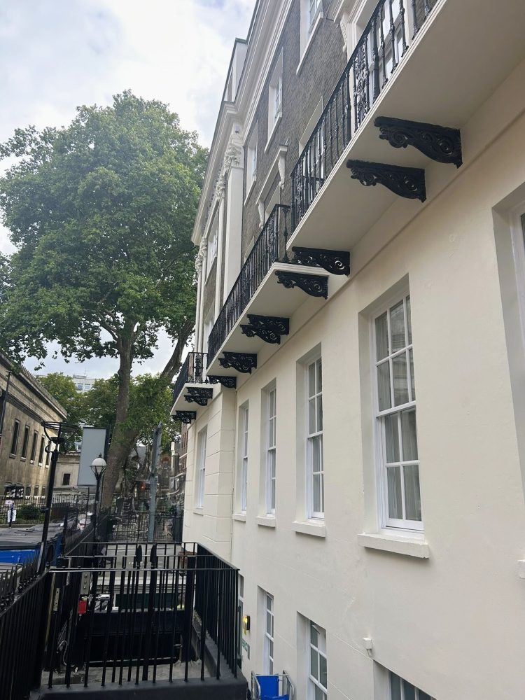 A close-up view of a Hilton Euston’s historic façade showcasing decorative balconies and iron railings, with trees in the background.