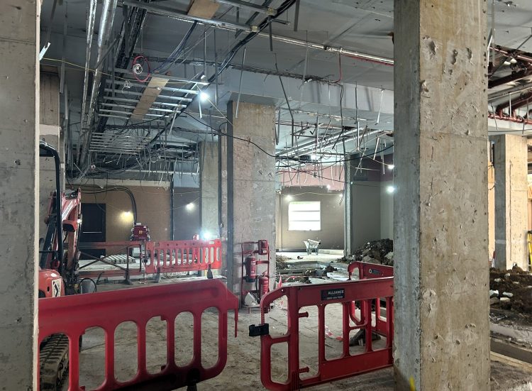 Construction site interior featuring exposed beams, scaffolding, red barriers, machinery, and workers amidst ongoing renovations.