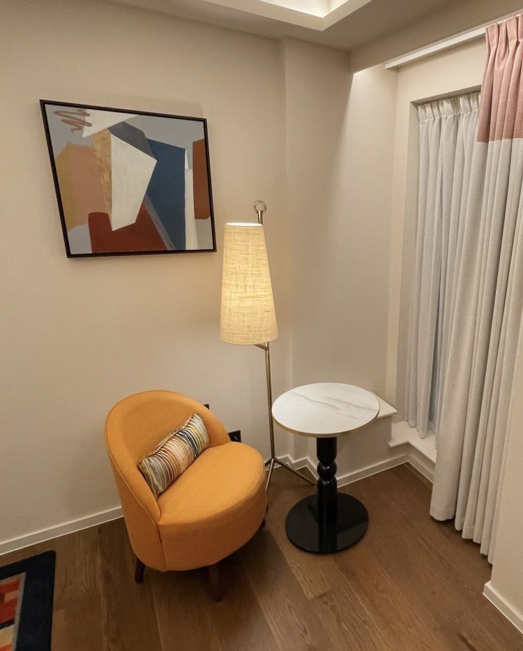A cozy corner of the new guest bedroom, featuring a vibrant orange chair with a striped pillow, a tall lampshade, and a round marble-top side table. An abstract painting adorns the wall, enhancing the warm, inviting atmosphere.