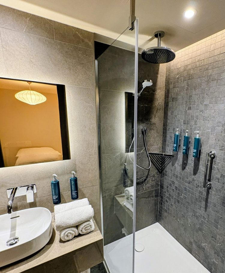 Modern bathroom with a glass shower, rain showerhead, and wall-mounted toiletries. The  sink with towels and a mirror reflecting a cozy bedroom.