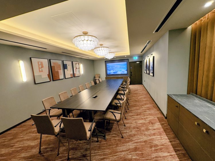 Modern conference room with a long, dark table and beige chairs. Art on walls, soft lighting from decorative ceiling lamps, and a screen at the far end. Cozy ambiance.