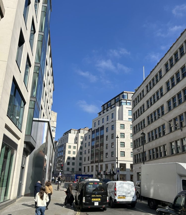 A city street scene with modern office buildings, several vehicles including a black taxi and white vans, and people walking on the sidewalk under a clear blue sky.