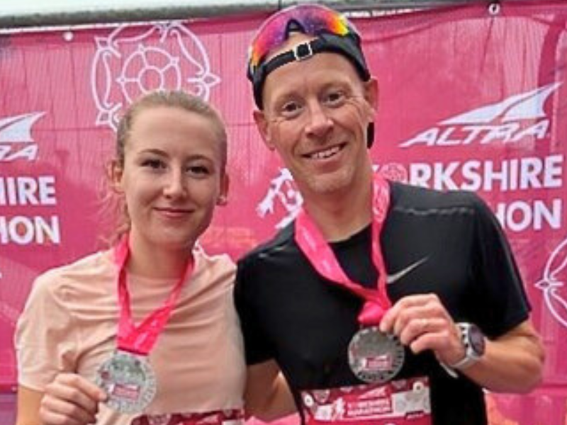 Sophie and her Dad proudly showing off their medals in front of a bright pink background.