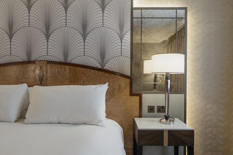 Modern hotel room with a plush bed, white pillows, and an art deco headboard. A sleek bedside lamp emits soft light beside a mirrored wall.
