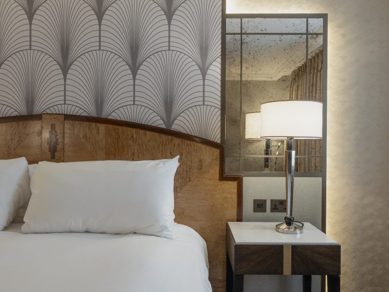 Modern hotel room with a plush bed, white pillows, and an art deco headboard. A sleek bedside lamp emits soft light beside a mirrored wall.