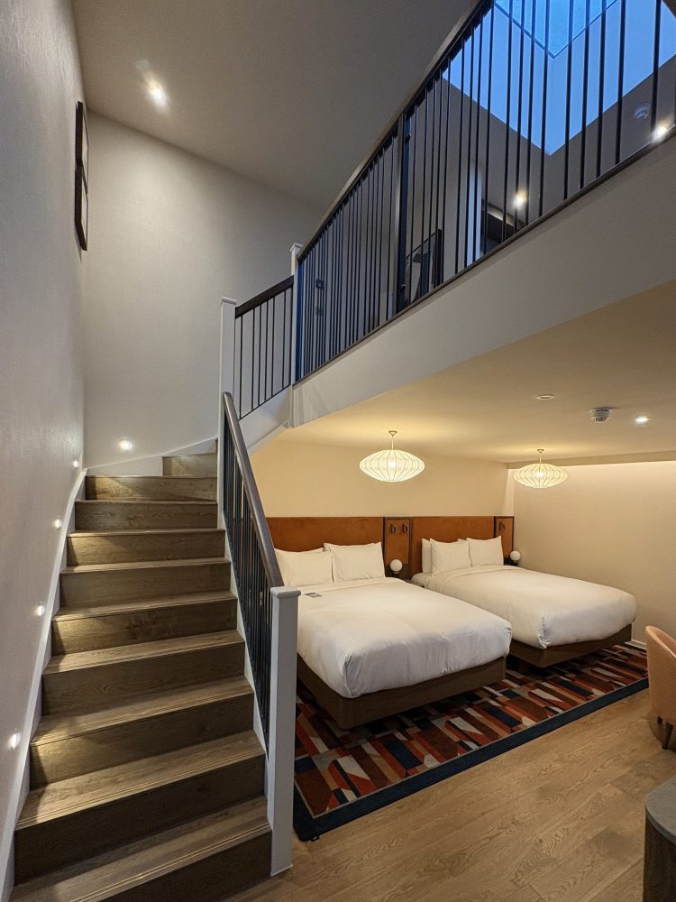 Modern hotel room featuring two double beds, a staircase, and stylish lighting in an open, airy layout.