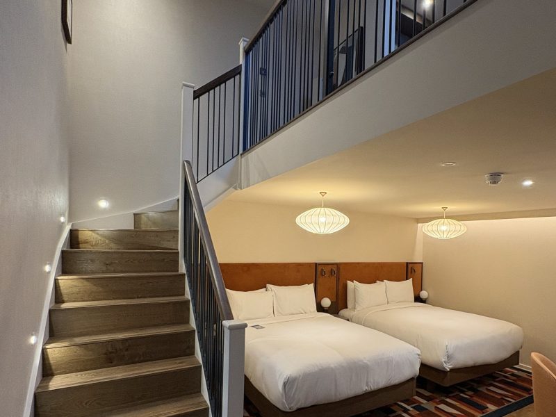 Modern hotel room featuring two double beds, a staircase, and stylish lighting in an open, airy layout.