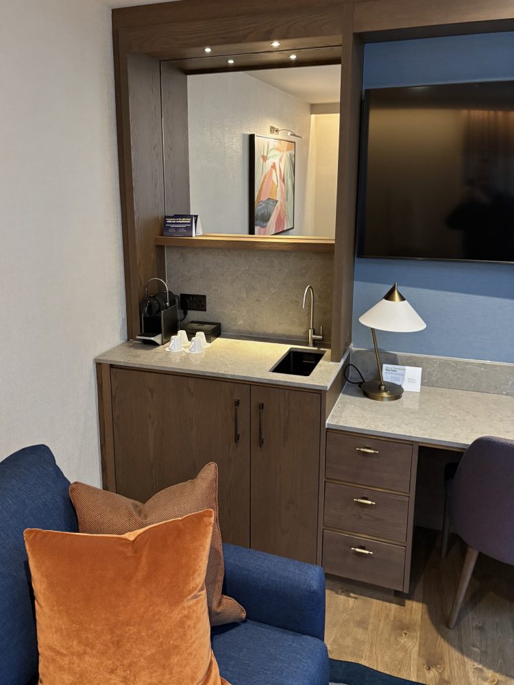 Cosy hotel room corner featuring a blue couch with orange pillows, a minimalist kitchenette, wall art, and a television.