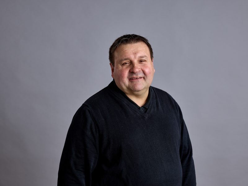 Photo of Justin wearing a black jumper smiling at the camera. He's in front of a grey background.