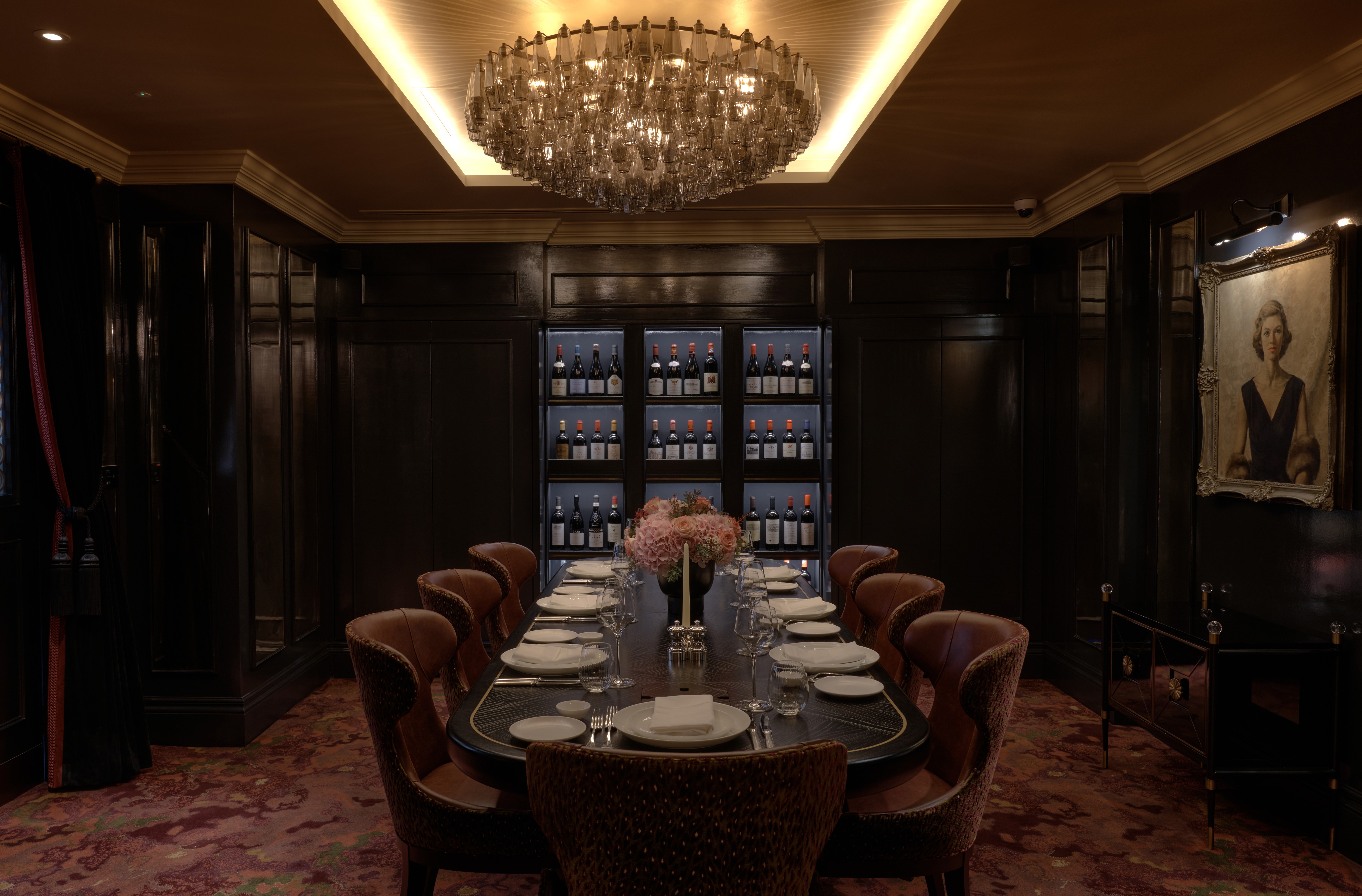 Elegant dining room with a long table set for 12, adorned with pink flowers. A chandelier hangs above, wine bottles line the dark wood walls, and a portrait is displayed.