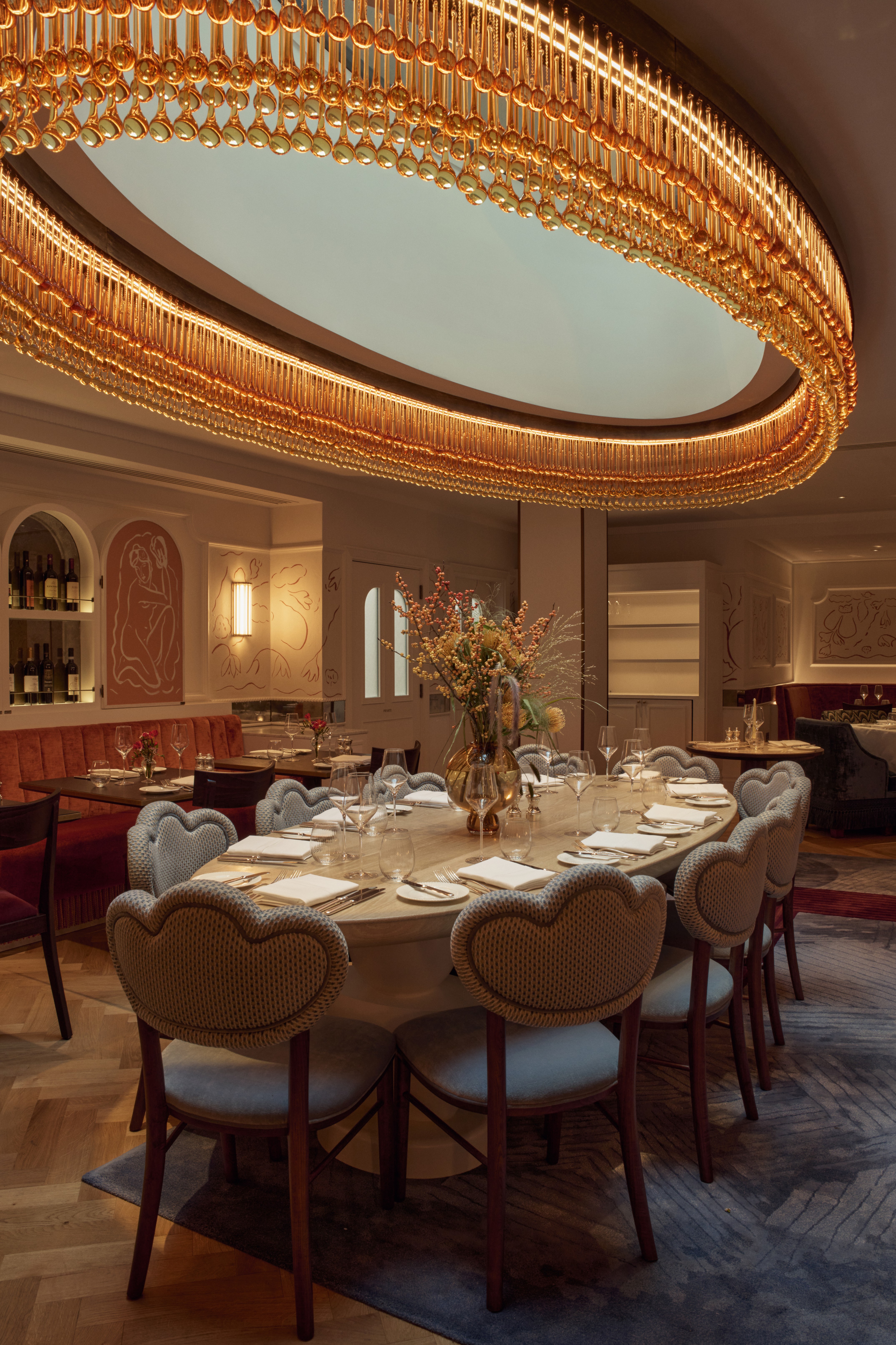 Elegant restaurant interior featuring a large oval table with scallop edge chairs, set for dining. A large golden oval chandelier adds warmth and luxury.