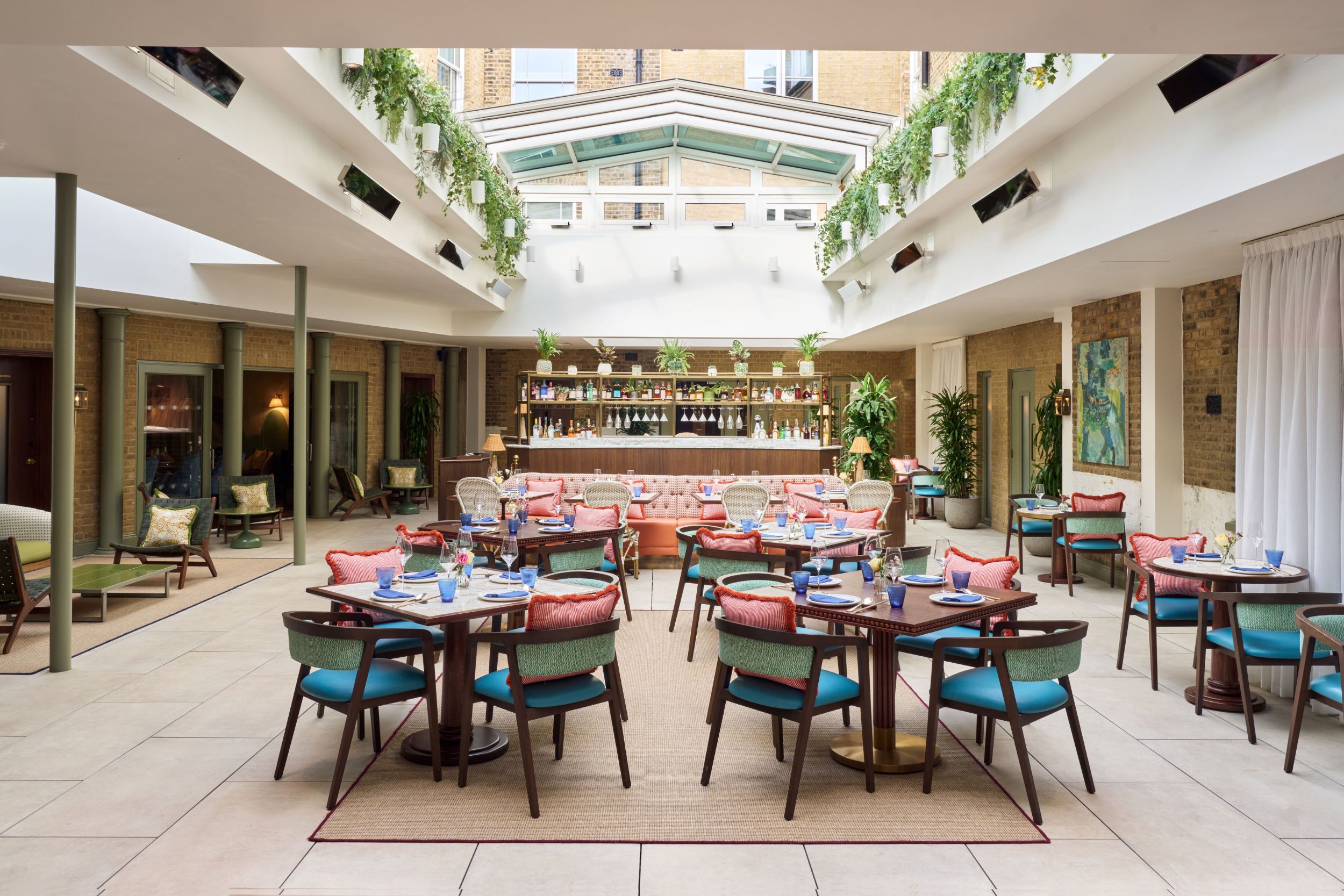 The Sloane Club - LILA covered terrace with the roof retracted. A pale stone tiled floor with teal and coral upholstered dark wood chairs and tables. A bar is in the background.