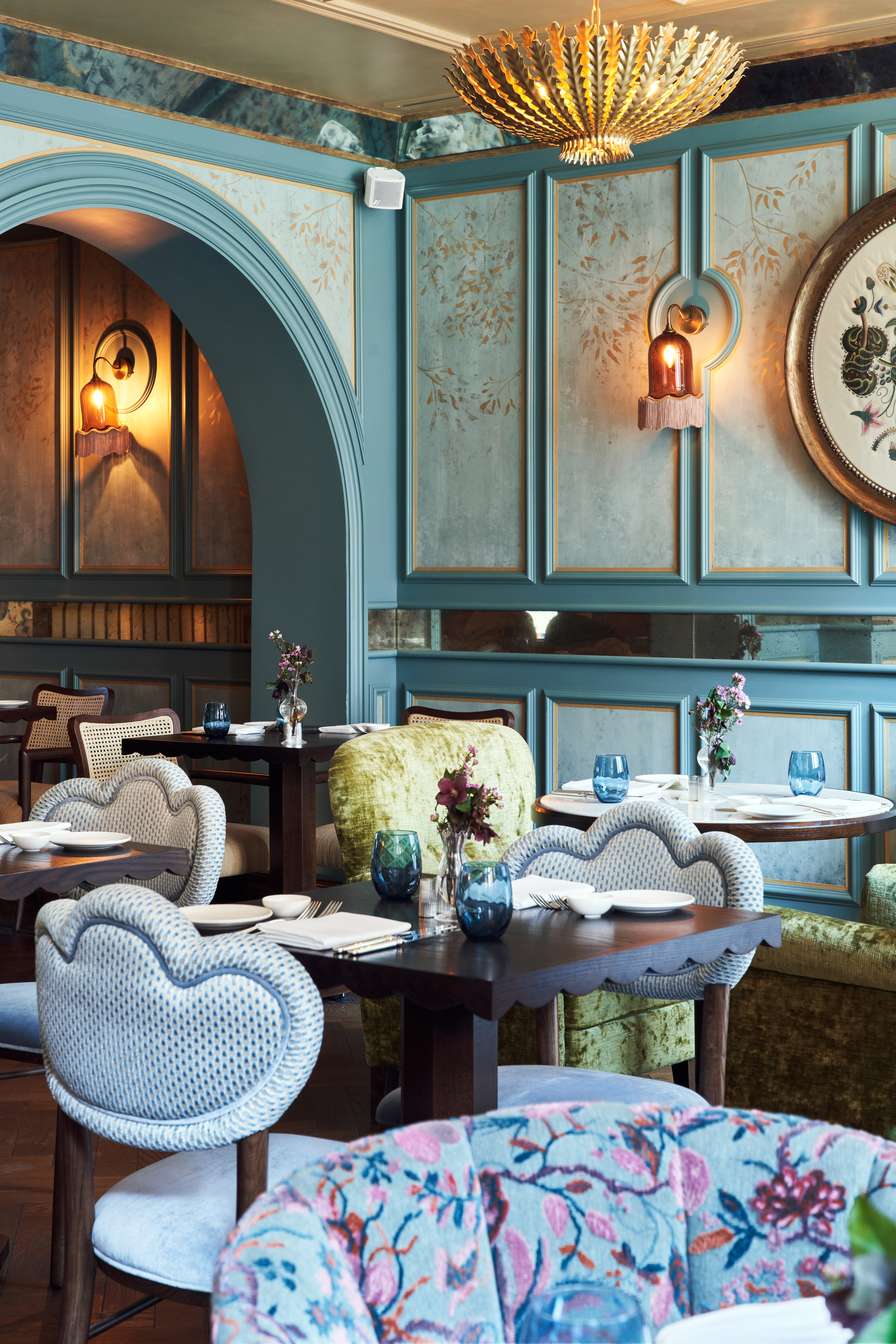 A luxurious dining space with decorative blue / teal panelled walls, feature gold pendant light, dark wood tables and different patterned / colour chairs in blues, greens and florals.
