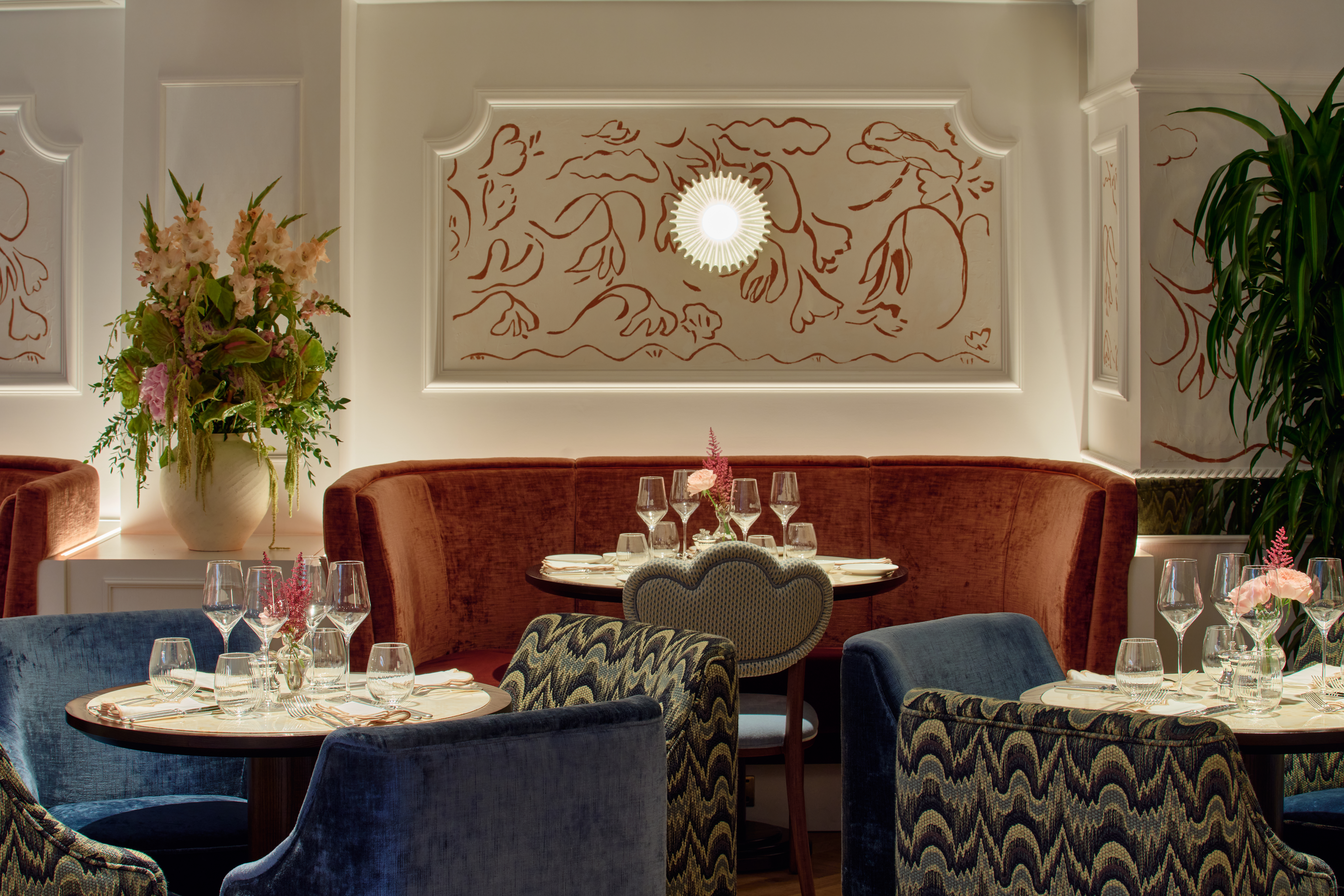 A luxurious dining space with light walls and decorative line illustration in terracotta. There is a deep terracotta bench seat against the wall and in the foreground are who circular tables with blue seats. There is a large floral display next to the bench seat area. 
