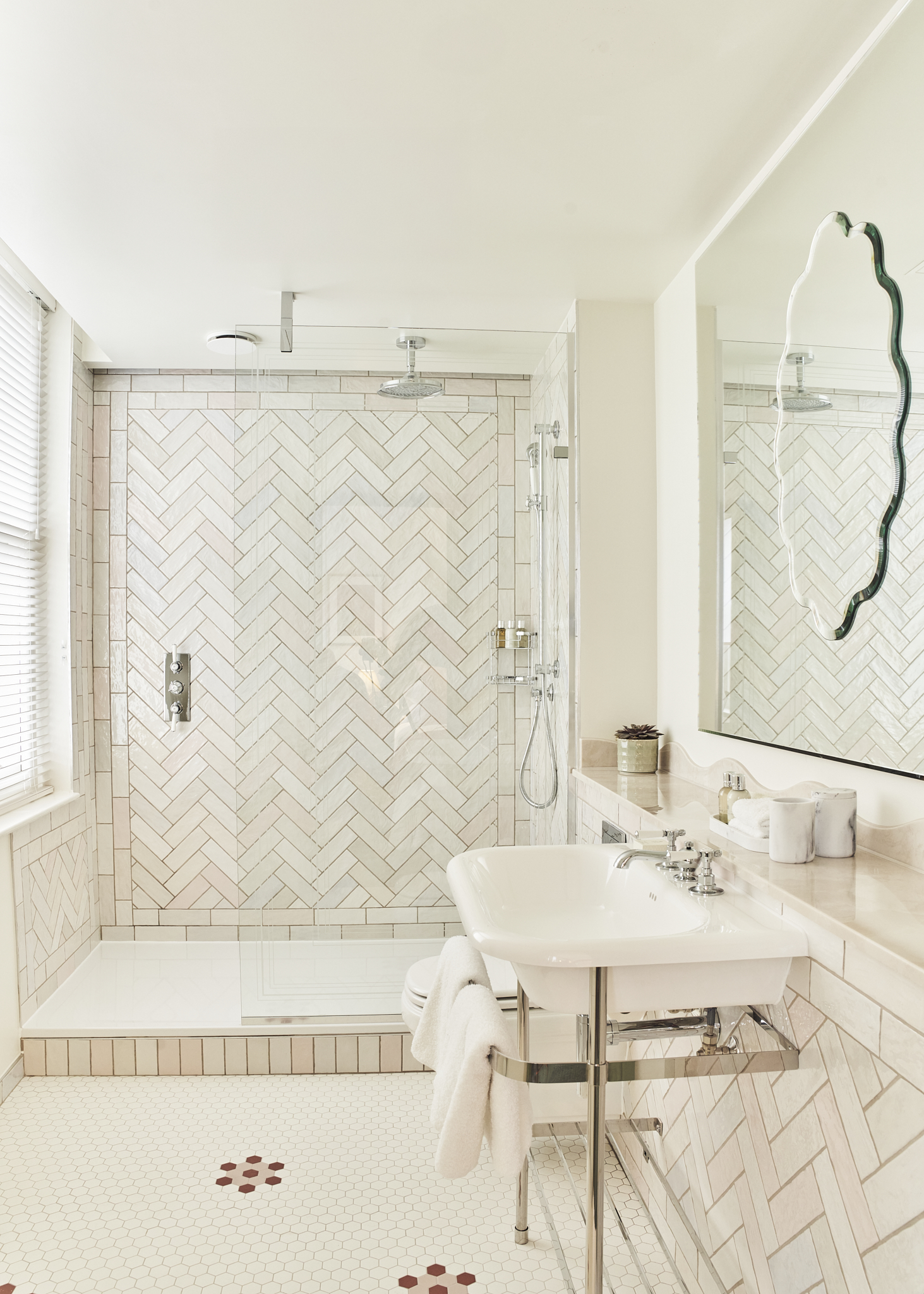 A spacious guest ensuite with large walk in shower, white sink and toilet. The floor tiles are small white hexagons with a pink / burgundy pattern in places. The wall tiles are brick shaped and laid in a chevron pattern - they are off-white / blush.
