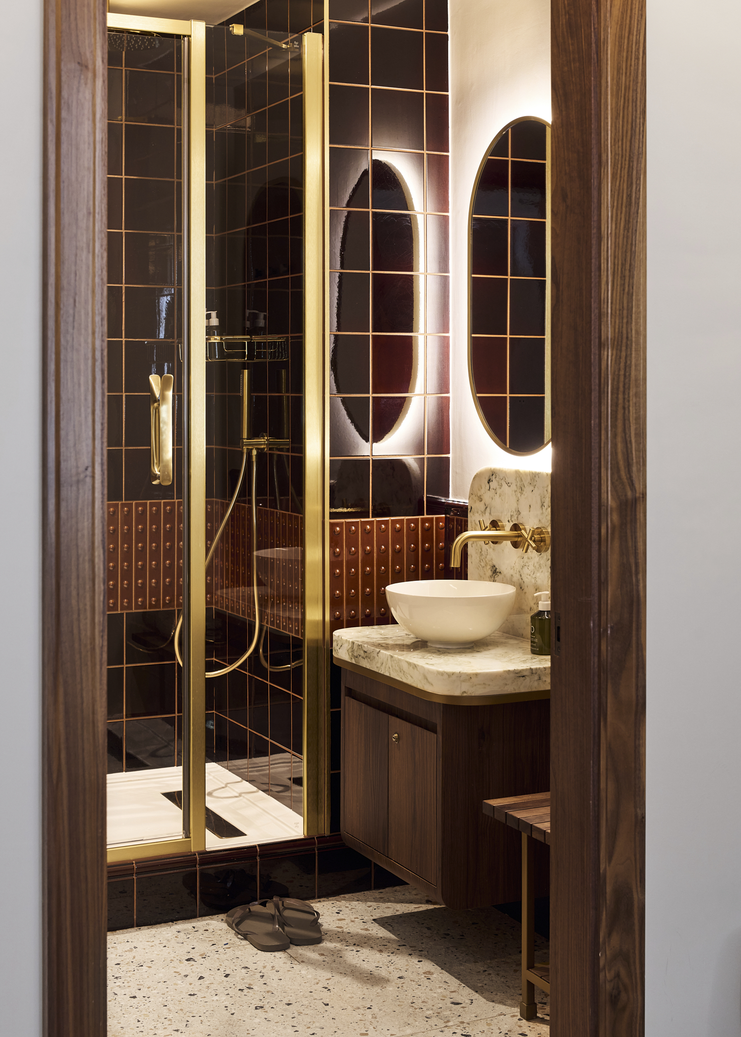The bathroom area in the wellness suite, featuring dark tiles and wooden vanity, a white bowl sink and gold hardware.
