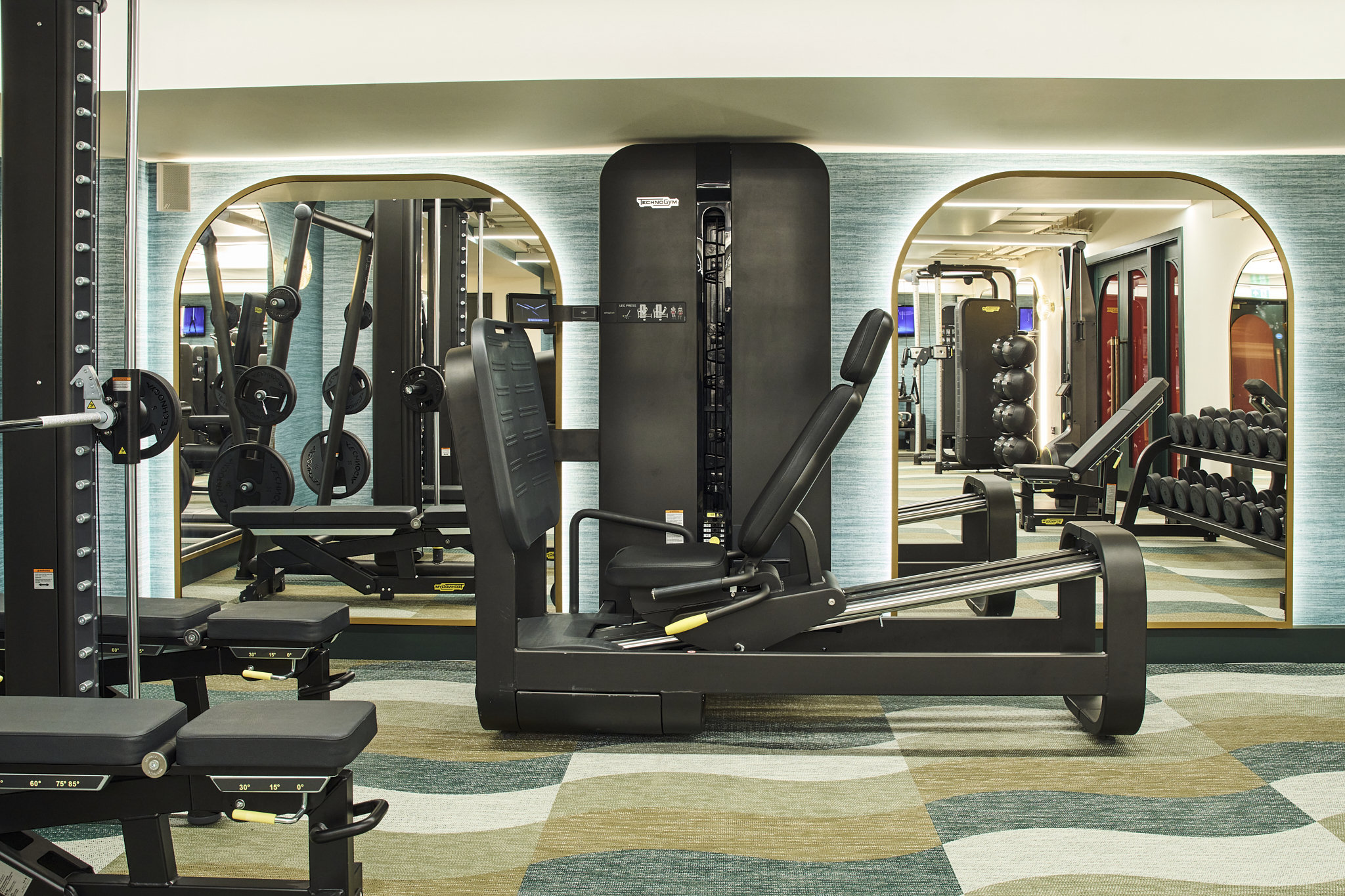 The gym area with teal, beige and cream wave patterned carpet, black gym equipment and large mirrors which are lit from behind,