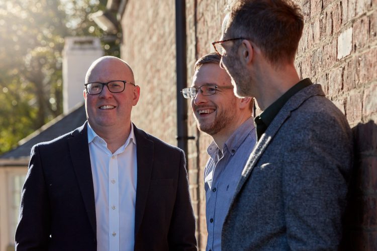 Peter Newman-Earp standing with Directors David Gardner and Nik Clarke outside on a sunny day with a red brick wall behind them