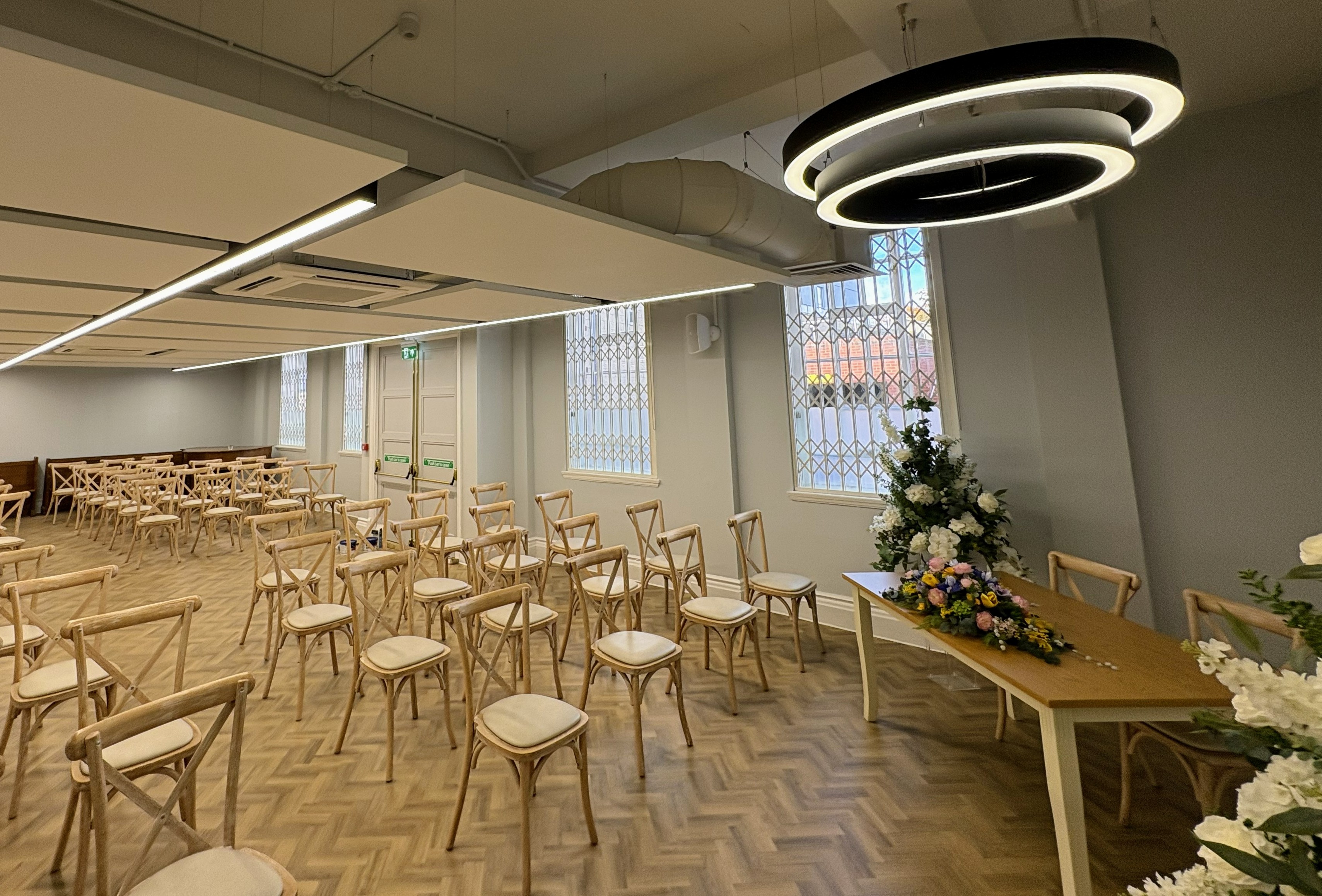 Large ceremony room with circular feature light at the front, over a table with a floral display. Chairs are set out ready for guests.